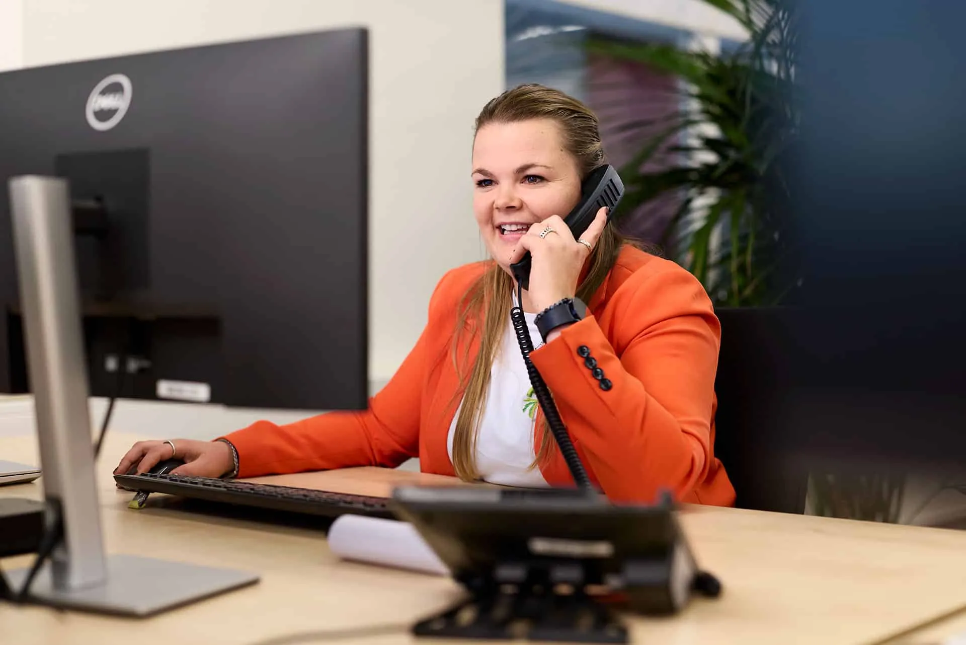 Customer service representative in an orange blazer speaks on a corded phone at her desk in an office setting.