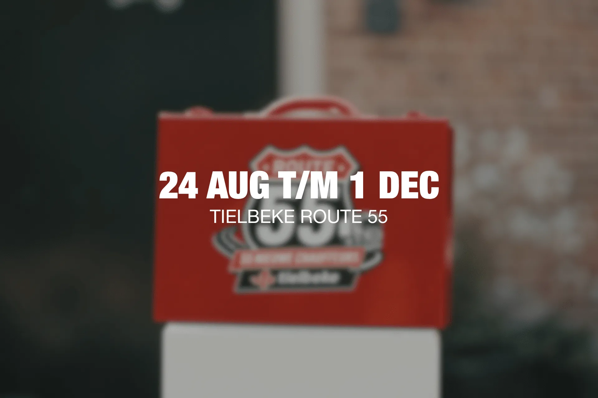 Red promotional bag with bold text showing dates 24 Aug to 1 Dec and location Tielbeke Route 55, blurred background marketing image.