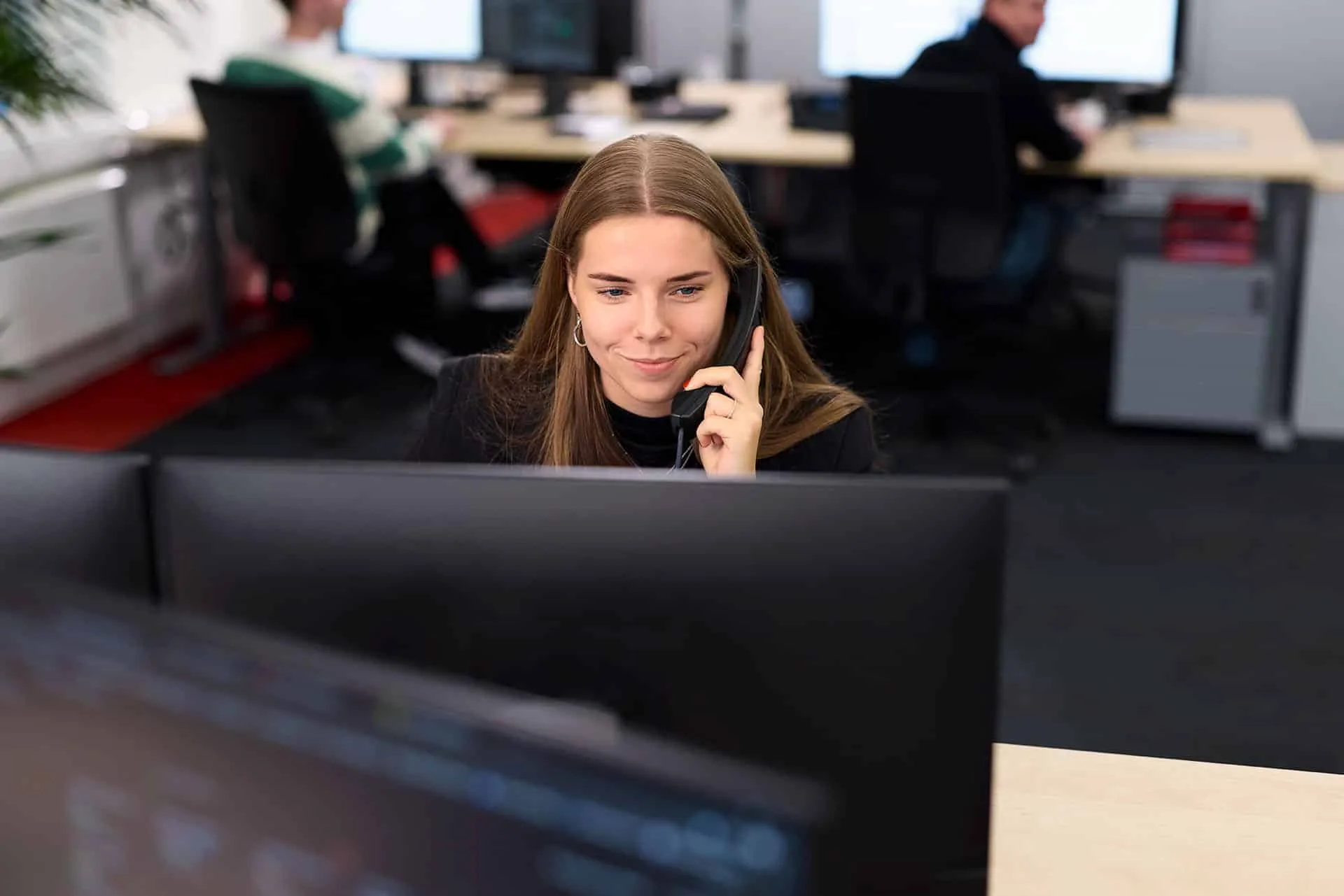 Woman wearing a headset works at a desk in a busy office, looking at a computer monitor.